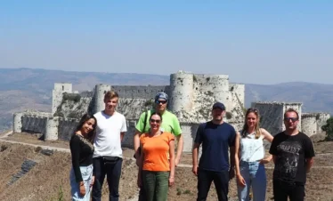 A group of six travelers in casual summer clothing stands on a hillside, posing in front of the massive stone walls and round towers of the Krak des Chevaliers crusader castle under a clear blue sky.