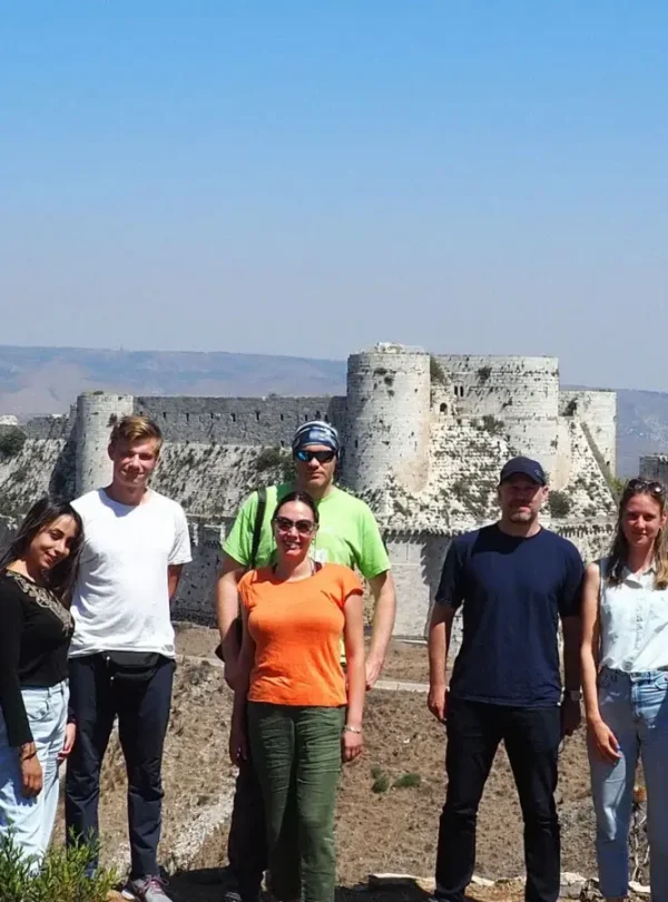 A group of six travelers in casual summer clothing stands on a hillside, posing in front of the massive stone walls and round towers of the Krak des Chevaliers crusader castle under a clear blue sky.