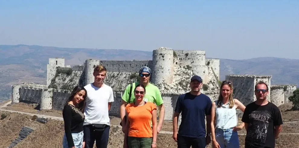 A group of six travelers in casual summer clothing stands on a hillside, posing in front of the massive stone walls and round towers of the Krak des Chevaliers crusader castle under a clear blue sky.