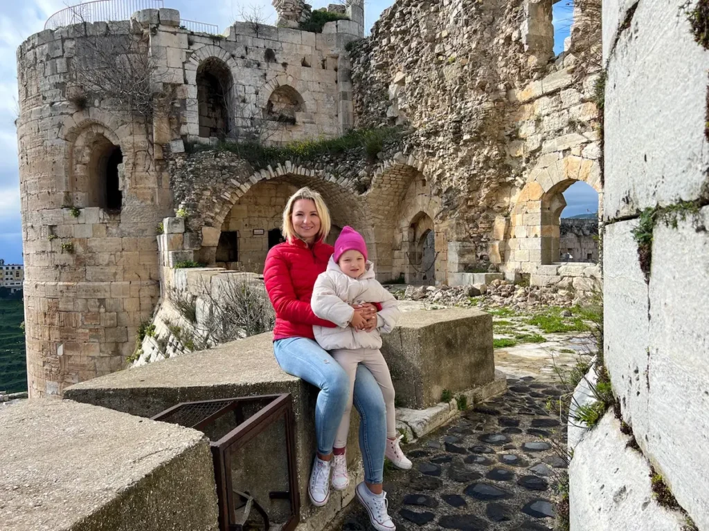 A woman in a red jacket and a young child in a pink hat sit together on a stone ledge within the ancient, weathered walls and arched passages of the Krak des Chevaliers castle.