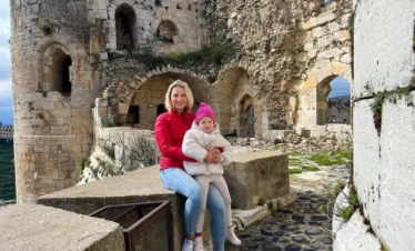 A woman in a red jacket and a young child in a pink hat sit together on a stone ledge within the ancient, weathered walls and arched passages of the Krak des Chevaliers castle.
