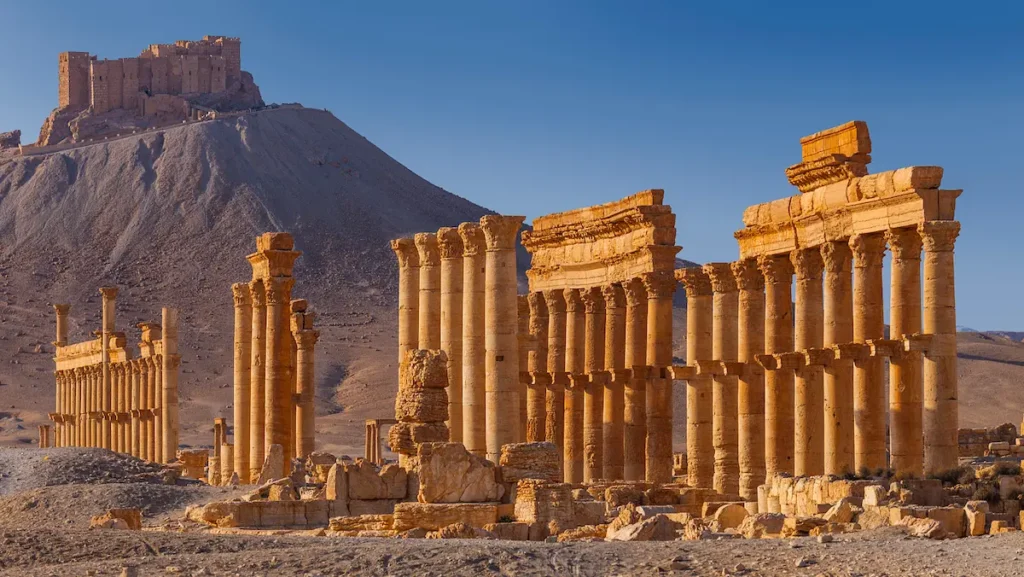 Golden-hued Roman columns and ruins of the ancient city of Palmyra in the foreground, with the hilltop Fakhr-al-Din al-Maani Castle (Qala'at Shirkuh) overlooking the site in the background.