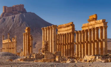 Golden-hued Roman columns and ruins of the ancient city of Palmyra in the foreground, with the hilltop Fakhr-al-Din al-Maani Castle (Qala'at Shirkuh) overlooking the site in the background.