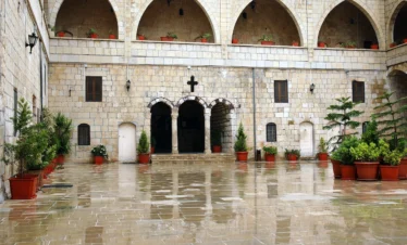 A wide, sunlit view of a wet stone courtyard at the Saint George Monastery, featuring large arched openings on the upper levels, potted green plants, and a central entrance with three smaller arches under a stone cross.
