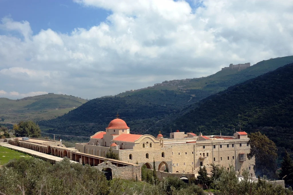 A high-angle view of the large stone Monastery of Saint George Al-Humaira with its distinct red domes, nestled in a lush green valley with the silhouette of Krak des Chevaliers castle on the distant hilltop under a cloudy sky.