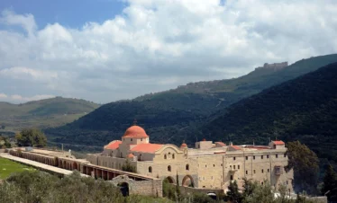 A high-angle view of the large stone Monastery of Saint George Al-Humaira with its distinct red domes, nestled in a lush green valley with the silhouette of Krak des Chevaliers castle on the distant hilltop under a cloudy sky.