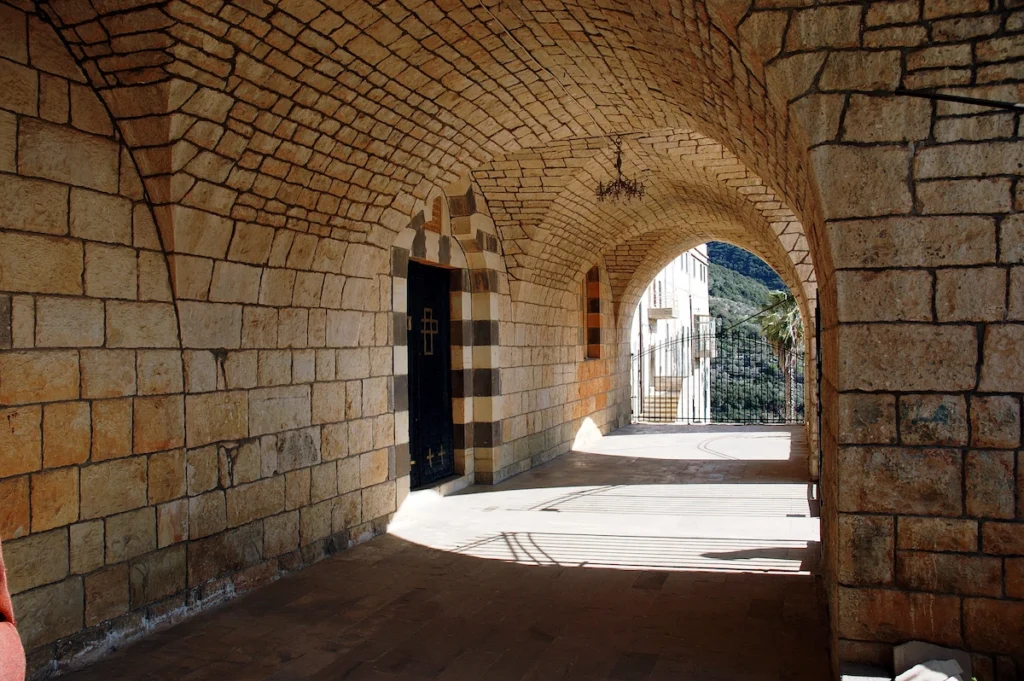 A wide stone-paved corridor within the Saint George Monastery, featuring large limestone walls and a vaulted ceiling that leads toward an arched exit overlooking a green mountain valley.