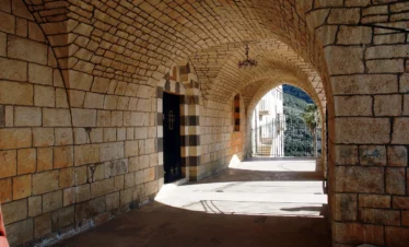 A wide stone-paved corridor within the Saint George Monastery, featuring large limestone walls and a vaulted ceiling that leads toward an arched exit overlooking a green mountain valley.