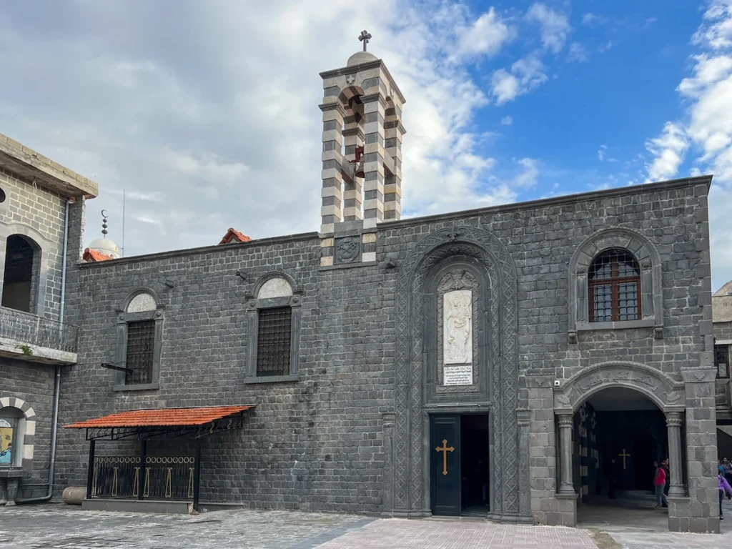 The exterior of an ancient church built from dark grey basalt stone, featuring a striped black-and-white bell tower, arched windows, and a marble relief above the main entrance under a blue sky.