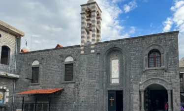 The exterior of an ancient church built from dark grey basalt stone, featuring a striped black-and-white bell tower, arched windows, and a marble relief above the main entrance under a blue sky.
