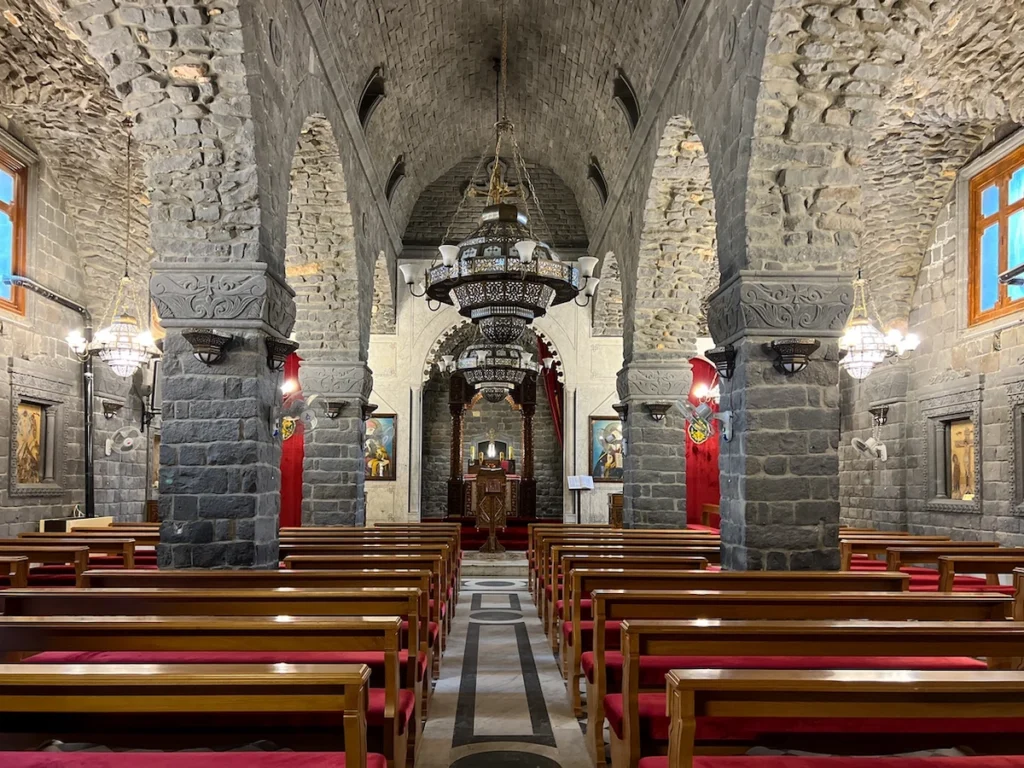 The interior of a historic church featuring massive grey basalt stone pillars and arches, wooden pews with red cushions, and large ornate metal chandeliers hanging from a vaulted ceiling leading toward a distant altar.
