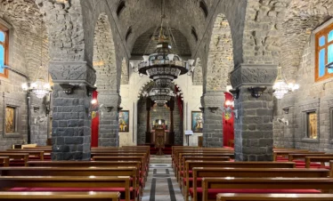 The interior of a historic church featuring massive grey basalt stone pillars and arches, wooden pews with red cushions, and large ornate metal chandeliers hanging from a vaulted ceiling leading toward a distant altar.