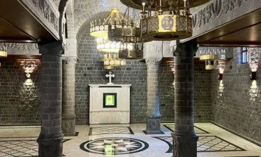 A wide interior view of a lower church hall with polished marble floors featuring green and white geometric patterns, massive grey basalt stone pillars, and large ornate gold chandeliers hanging from a vaulted ceiling.