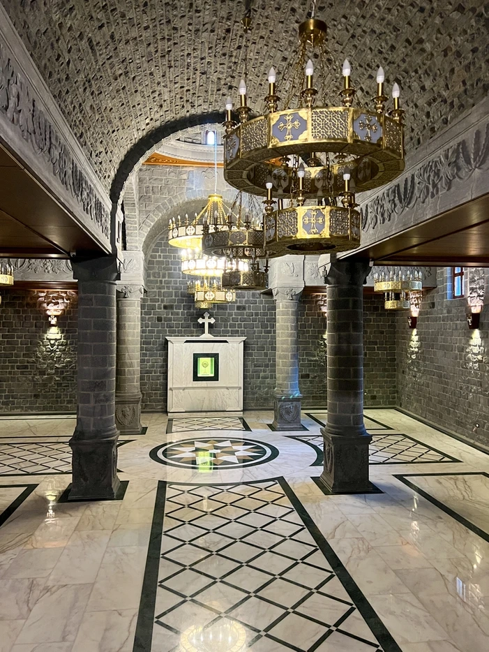 A wide interior view of a lower church hall with polished marble floors featuring green and white geometric patterns, massive grey basalt stone pillars, and large ornate gold chandeliers hanging from a vaulted ceiling.