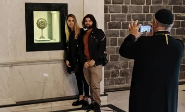 A man wearing a traditional red tarbush and grey robe sits in a wood-paneled café, holding a sword and reading from a small book to a group of men sitting in the background with hookahs.