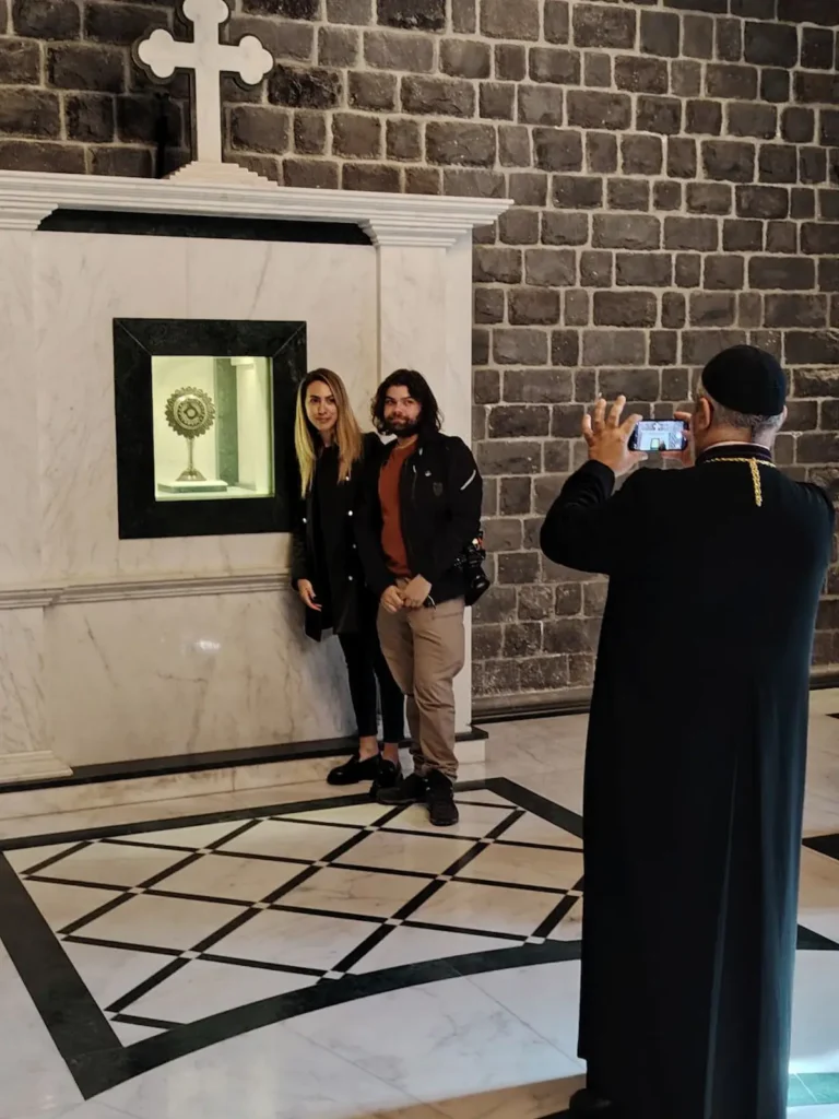 A man wearing a traditional red tarbush and grey robe sits in a wood-paneled café, holding a sword and reading from a small book to a group of men sitting in the background with hookahs.