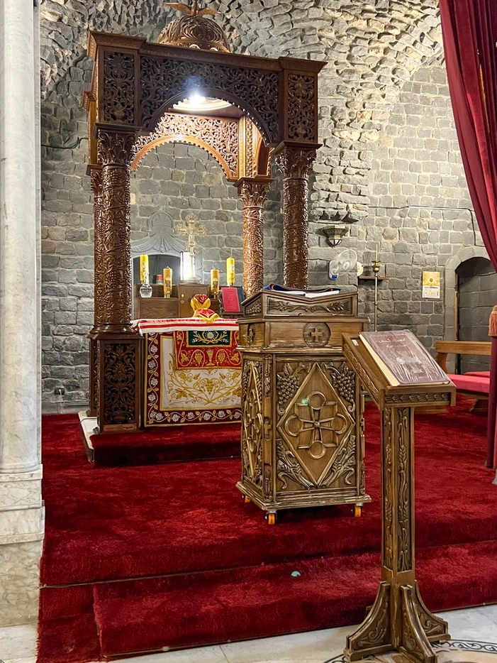 The ornate wooden altar of an ancient church, featuring four tall, intricately carved pillars supporting a decorative canopy over a red-carpeted platform, set against a backdrop of historic grey basalt stone walls.