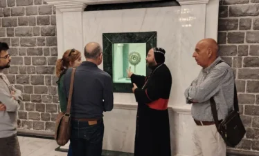 A priest in black and red robes gestures toward a golden reliquary displayed in a marble wall niche while a group of four travelers listens intently inside a church with dark basalt stone walls and a white marble floor.