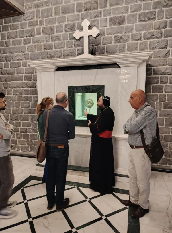 A priest in black and red robes gestures toward a golden reliquary displayed in a marble wall niche while a group of four travelers listens intently inside a church with dark basalt stone walls and a white marble floor.
