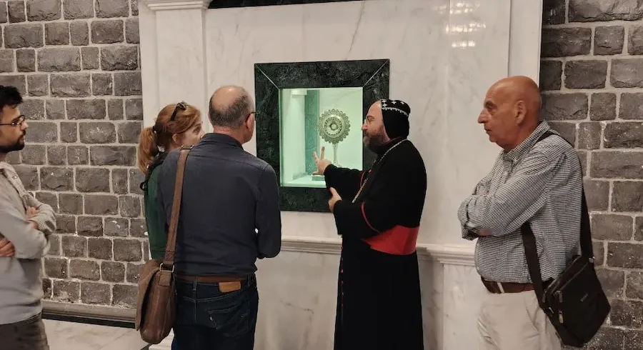 A priest in black and red robes gestures toward a golden reliquary displayed in a marble wall niche while a group of four travelers listens intently inside a church with dark basalt stone walls and a white marble floor.