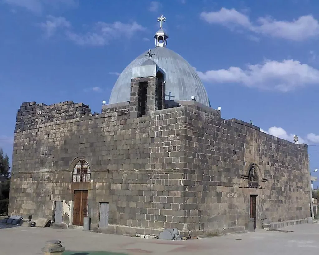 An exterior view of the ancient St. George’s Church in Izra’a, Syria, constructed from dark black basalt blocks with a large silver dome topped with a cross against a blue sky.