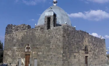 An exterior view of the ancient St. George’s Church in Izra’a, Syria, constructed from dark black basalt blocks with a large silver dome topped with a cross against a blue sky.