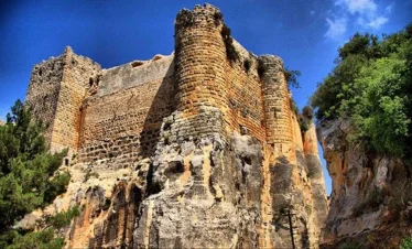 A low-angle view of the massive stone fortifications of the Citadel of Salah Ed-Din, featuring towering rounded bastions built directly into a high limestone cliff under a deep blue sky.