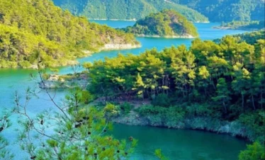 A high-angle view of a serene turquoise lake winding through lush green pine-covered mountains, with a white leisure boat anchored in a small cove under a clear blue sky.