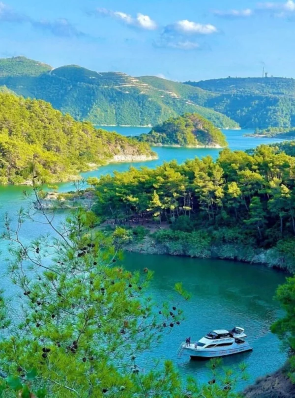 A high-angle view of a serene turquoise lake winding through lush green pine-covered mountains, with a white leisure boat anchored in a small cove under a clear blue sky.