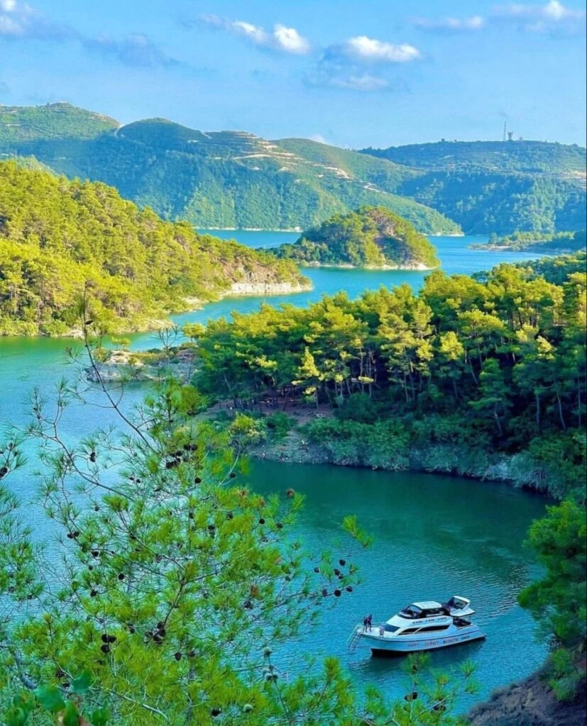 A high-angle view of a serene turquoise lake winding through lush green pine-covered mountains, with a white leisure boat anchored in a small cove under a clear blue sky.