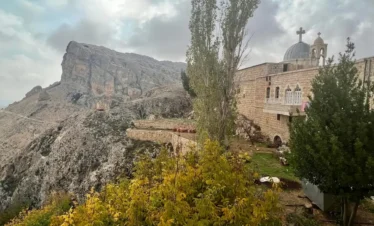 A view of the stone Saint Thecla Monastery built into a rugged, mountainous cliffside in Maaloula, Syria, featuring a domed chapel with a cross and yellow autumn foliage in the foreground.