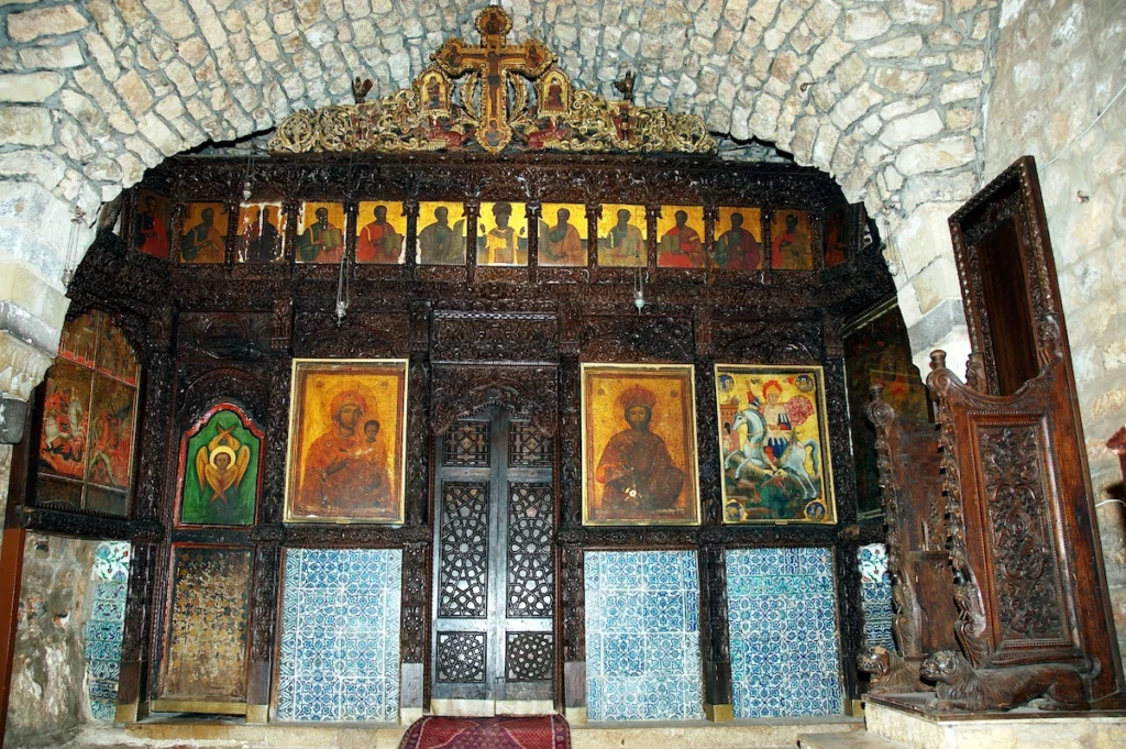 An intricately carved dark wood iconostasis in an ancient stone church, featuring gold-leaf icons of saints, a large central painting of the Virgin and Child, and blue decorative tiling along the base.
