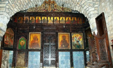 An intricately carved dark wood iconostasis in an ancient stone church, featuring gold-leaf icons of saints, a large central painting of the Virgin and Child, and blue decorative tiling along the base.