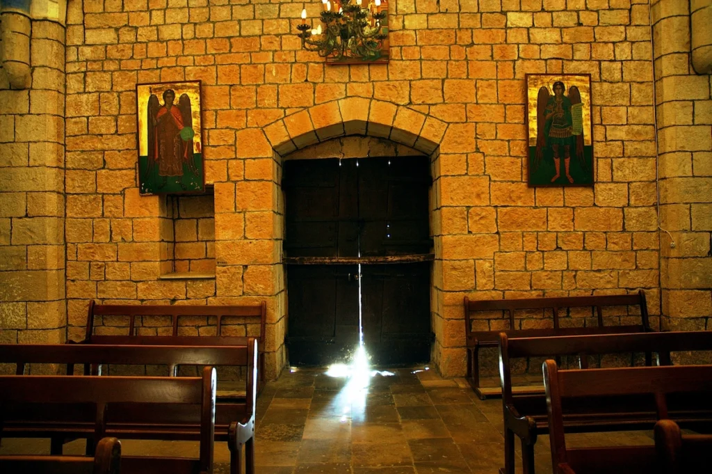 A soft beam of light streams through a crack in a heavy, dark wooden door inside an ancient stone church, illuminating the pews and golden-hued masonry walls adorned with icons of angels.