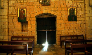 A soft beam of light streams through a crack in a heavy, dark wooden door inside an ancient stone church, illuminating the pews and golden-hued masonry walls adorned with icons of angels.