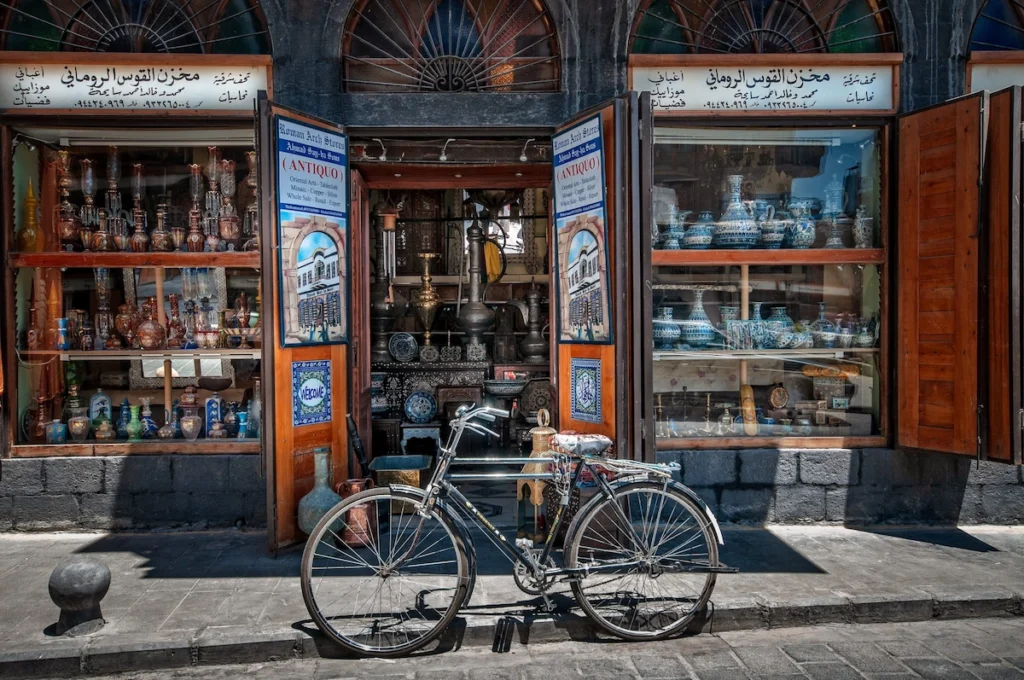 A classic black bicycle parked in front of an open-air antique shop in Old Damascus, with shelves displaying colorful mosaic glassware, copper vessels, and traditional Syrian handicrafts under stone arches.