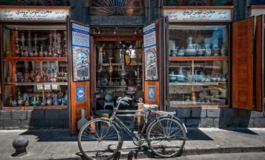 A classic black bicycle parked in front of an open-air antique shop in Old Damascus, with shelves displaying colorful mosaic glassware, copper vessels, and traditional Syrian handicrafts under stone arches.