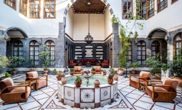 A wide-angle view of a luxurious open-air Damascene courtyard with a central marble fountain, intricate black-and-white stone walls (Ablaq), velvet armchairs, and lush green plants under a wooden ceiling.