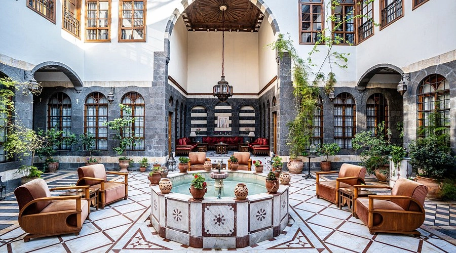 A wide-angle view of a luxurious open-air Damascene courtyard with a central marble fountain, intricate black-and-white stone walls (Ablaq), velvet armchairs, and lush green plants under a wooden ceiling.