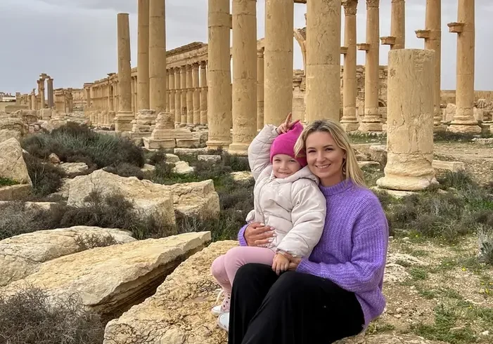 A woman in a purple sweater and a young child in a pink hat sit on a large ancient stone block, smiling together in front of the long rows of towering Roman columns at the Palmyra archaeological site under an overcast sky.
