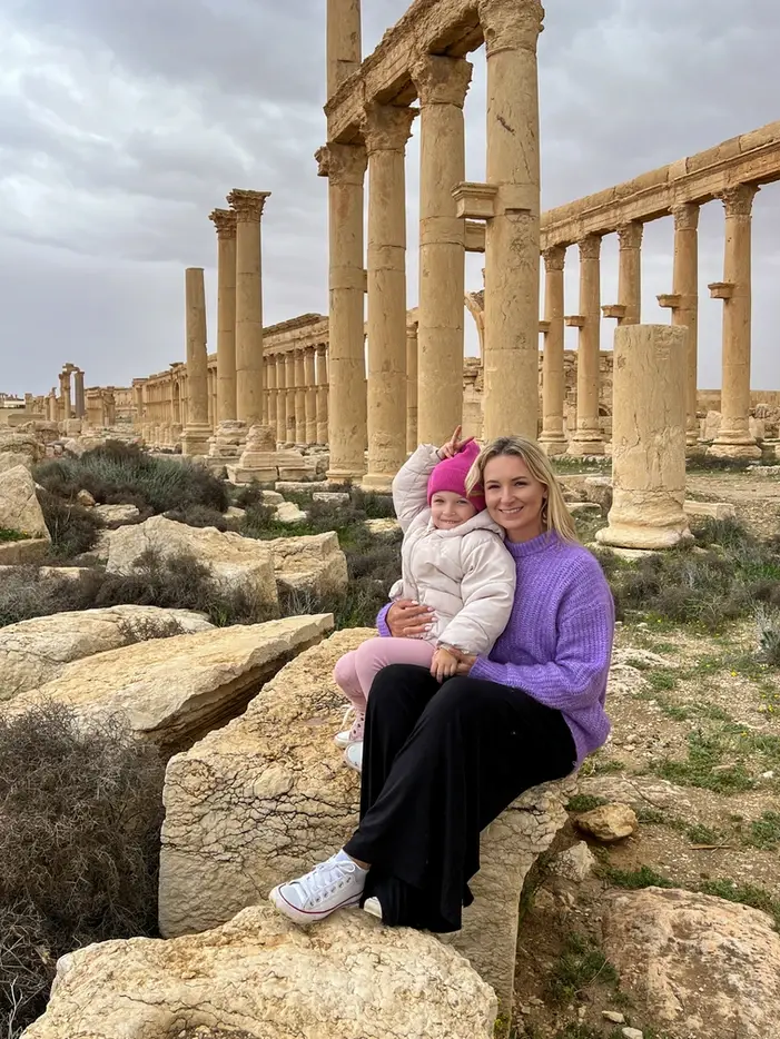 A woman in a purple sweater and a young child in a pink hat sit on a large ancient stone block, smiling together in front of the long rows of towering Roman columns at the Palmyra archaeological site under an overcast sky.