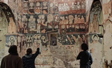 Three travelers stand on patterned carpets inside a stone chapel, looking up at a high wall completely covered in colorful medieval frescoes of saints and religious scenes.