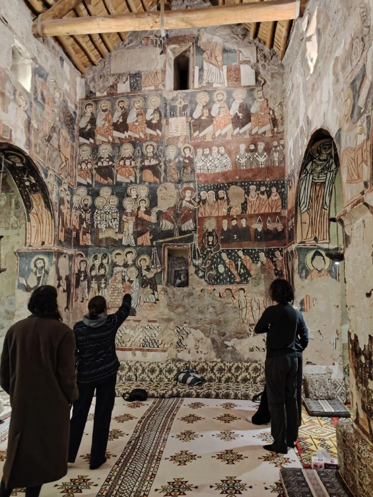 Three travelers stand on patterned carpets inside a stone chapel, looking up at a high wall completely covered in colorful medieval frescoes of saints and religious scenes.