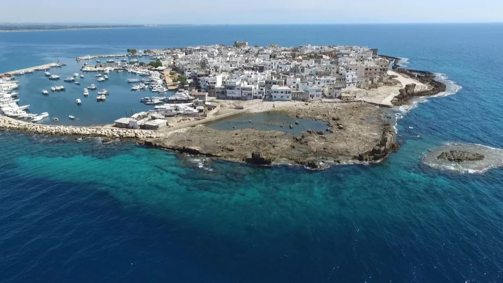 An aerial drone view of Arwad Island in the Mediterranean Sea, showing a densely packed town of white buildings, a small circular harbor filled with boats, and turquoise water along the rocky shoreline.