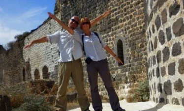 A man and a woman in casual travel attire pose playfully with their arms outstretched in front of the massive black basalt stone walls and arched windows of Margat Castle under a blue sky.
