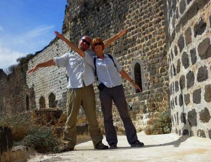 A man and a woman in casual travel attire pose playfully with their arms outstretched in front of the massive black basalt stone walls and arched windows of Margat Castle under a blue sky.