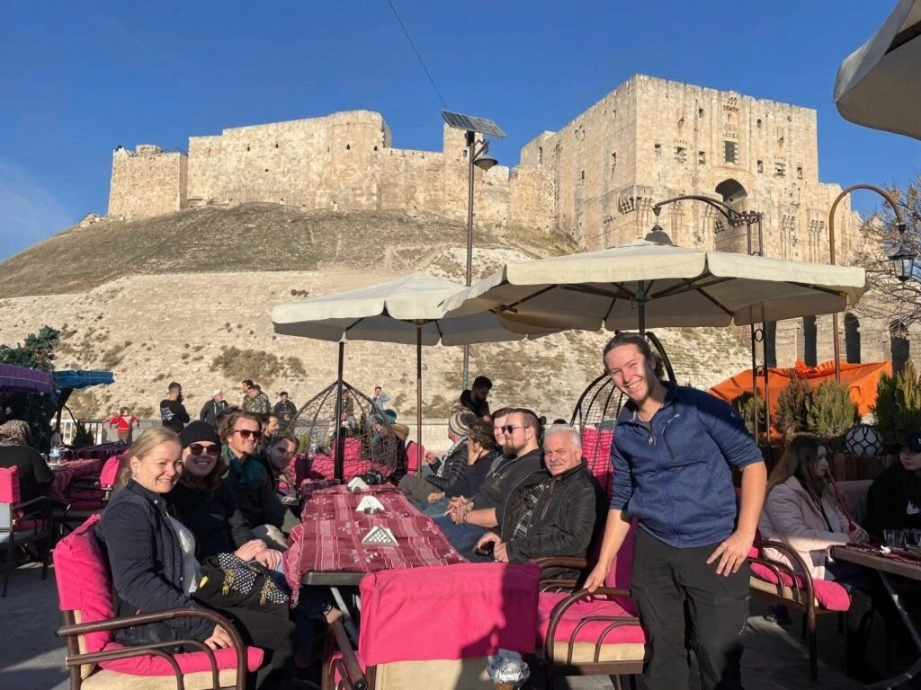 International travelers enjoying coffee at an outdoor cafe with a clear view of the ancient Aleppo Citadel in Syria.