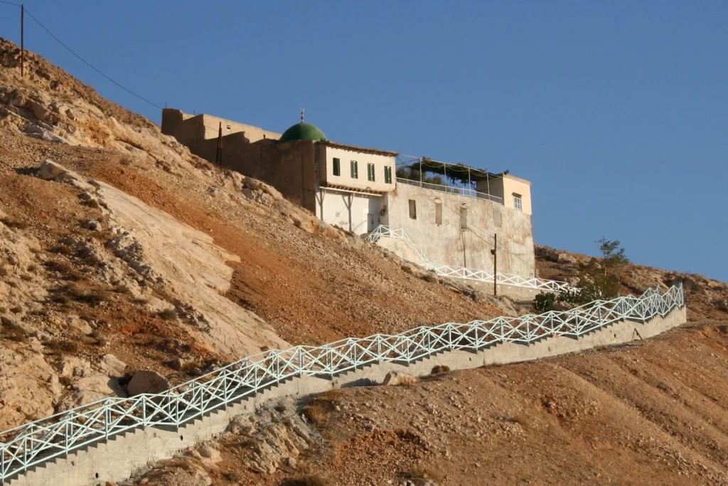 A view of the historic Arbain Mosque with its distinctive green dome, perched on the rocky, desert slopes of Mount Qasioun, featuring a long, zig-zagging staircase with a light blue railing leading up to the white building.