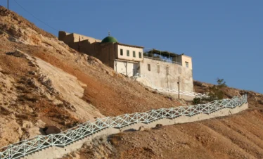 A view of the historic Arbain Mosque with its distinctive green dome, perched on the rocky, desert slopes of Mount Qasioun, featuring a long, zig-zagging staircase with a light blue railing leading up to the white building.