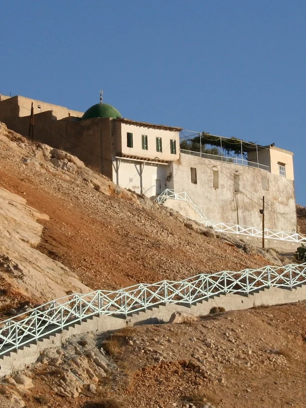 A view of the historic Arbain Mosque with its distinctive green dome, perched on the rocky, desert slopes of Mount Qasioun, featuring a long, zig-zagging staircase with a light blue railing leading up to the white building.